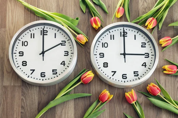 Two clocks, one showing two o\'clock, the other showing three o\'clock. Tulips around the second one. Time change symbol. Daylight saving time. Moving the hands forward from 2 a.m. to 3 a.m.