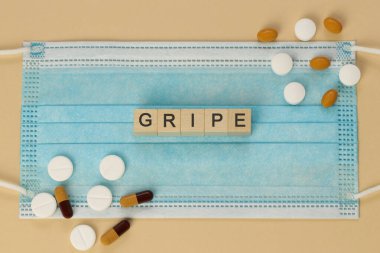 Gripe - influenza in spanish and portuguese, spelled with wooden cubes on a surgical mask, surrounded by pills and capsules. clipart