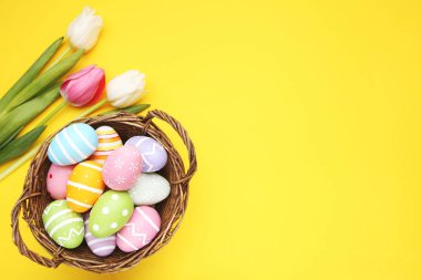 Colorful eggs in basket and flowers of tulips on yellow background. Easter day concept