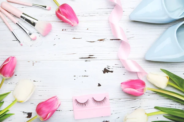 Flowers of tulips and makeup brushes, eyelashes, ribbon, blue high heeled shoes on white wooden background