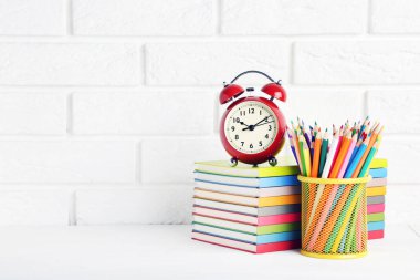 Renkli not defterlerindeki kırmızı alarm saati ve beyaz tuğla duvarlı arka plandaki kalemlerle dolu sepet.