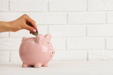 Beyaz tuğla duvar arka planında kumbaraya bozuk para atan bir kadın.