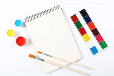 Fırçalarla renkli guaj boyaları ve beyaz ahşap masada boş kağıtlar.