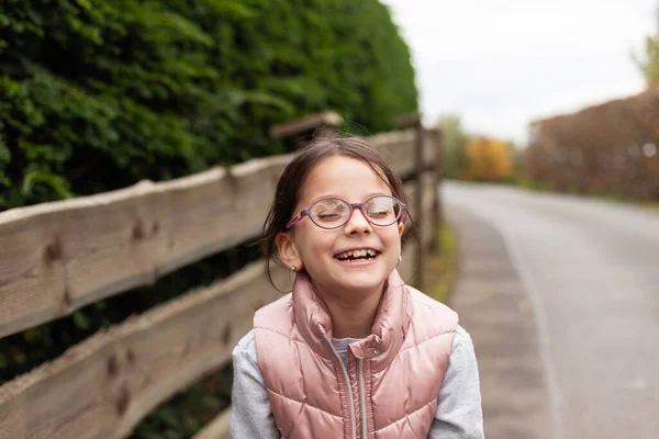 stock image Little beautiful cute girl in glasses laughing near a wooden fence. Village child in a vest on a walk in the countryside