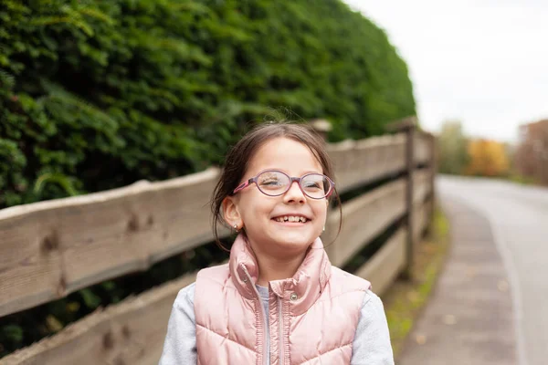 stock image Little beautiful cute girl in glasses laughing near a wooden fence. Village child in a vest on a walk in the countryside