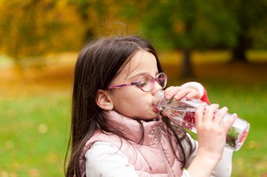 Uzun saçlı ve gözlüklü tatlı kız bulanık bir sonbahar parkında plastik şişeden su içiyor.