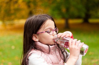 Uzun saçlı ve gözlüklü tatlı kız bulanık bir sonbahar parkında plastik şişeden su içiyor.