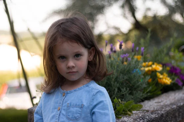 一个穿着斜纹棉布连衣裙的小女孩站在明亮的花朵旁边模糊的背景上 — 图库照片