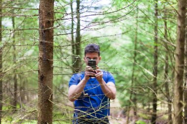  Genç bir adam ormanda duruyor, elinde bir kamera ve kendi fotoğrafını çekiyor. Etrafı gür ağaçlar ve yeşilliklerle çevrili. Güneş ışığı yaprakların arasından süzülüyor. Huzurlu ve huzurlu bir atmosfer yaratıyor..
