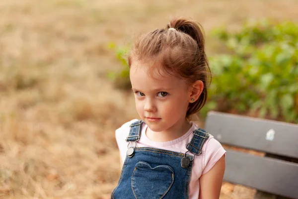 一个漂亮的金发碧眼的小女孩穿着斜纹棉布工作服坐在公园的长椅上 — 图库照片