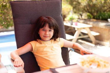 Küçük güzel gülen kız, sıcak bir sonbahar gününde arka bahçede bahçe sandalyesinde oturuyor.