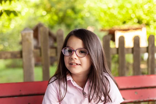 かわいい女の子の肖像画眼鏡をかけてベンチに座って — ストック写真