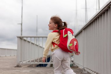 Sırtında kırmızı bir sırt çantası olan genç bir kız köprü boyunca yürüyor.