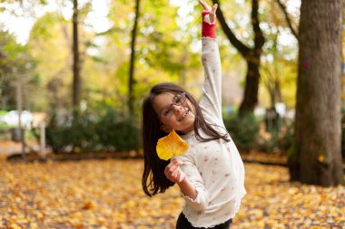 Sonbahar parkında elinde sarı bir yaprakla küçük bir kız.
