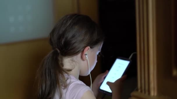 一个带着尾巴的小女孩在带耳机的智能手机上玩耍 — 图库视频影像