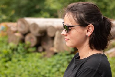 Bulanık doğal bir arka planda güneş gözlüklü ve ağaç gövdeleri kesen güzel bir kızın yan görüntüsü.