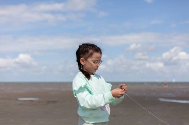 Little girl in clothes stands with a kite string on the beach clipart