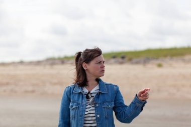 Cute girl in a denim jacket stands and looks into the distance on the beach clipart
