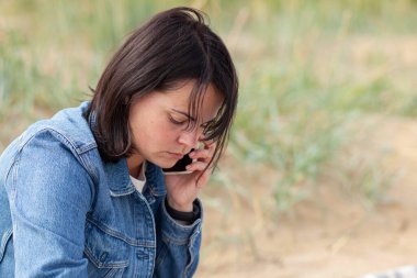 Koyu renk ceketli, güzel, siyah saçlı bir kız, ne yazık ki sahilde, deniz kenarında telefonla konuşuyor.