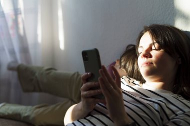 Genç bir kadın kanepede uzanıyor ve evde cep telefonu kullanıyor.