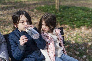Güneşli bir günde parkta plastik bir şişeden su içen iki kız.