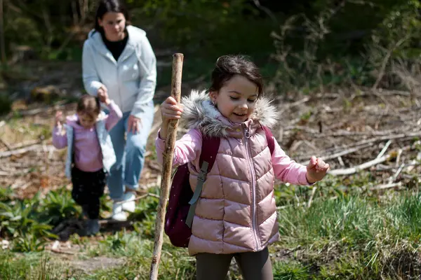 stock image A little girl with a stick and a staff walks through the forest. Mom and sister in the background