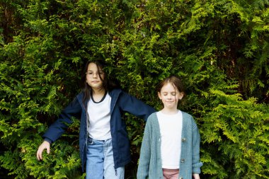 İki küçük kız kardeş yeşil bir çitin arkasında duruyor.