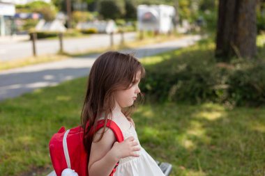 Kırmızı sırt çantalı küçük bir kız parkta bir bankta oturuyor.
