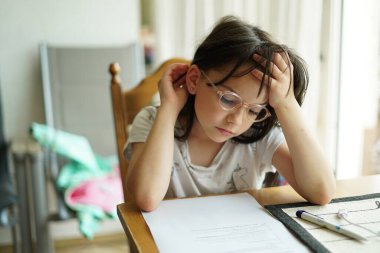 Gözlüklü küçük bir kız masada otururken ve ödevini yaparken ne yazık ki başını eline dayadı..
