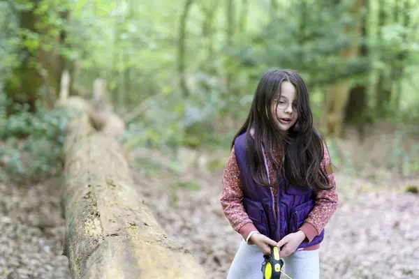 stock image A small beautiful smiling long-haired girl in glasses and a vest stands next to the trunk of a fallen tree in the forest