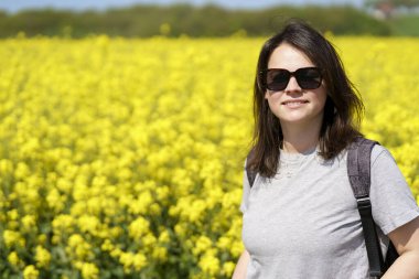 Güneş gözlüklü genç bir kadın çiçek açan kolza tohumu tarlasında duruyor.