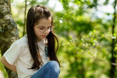 Portrait of a little girl in glasses sitting on a tree branch clipart