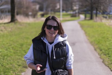 Güneş gözlüklü, yelek ve kapüşonlu tatlı siyah saçlı bir kız bahar parkında gülümsüyor.