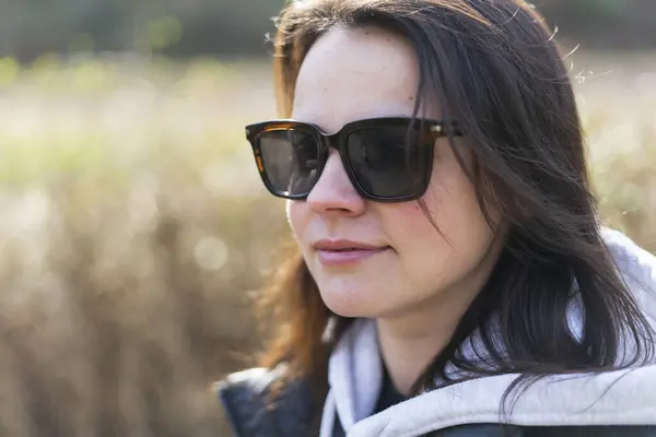 stock image A cute dark-haired girl in sunglasses, a vest and a hoodie stands smiling in a spring park