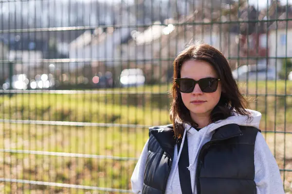 stock image A cute dark-haired girl in sunglasses, a vest and a hoodie stands smiling in a spring park in front of the fence