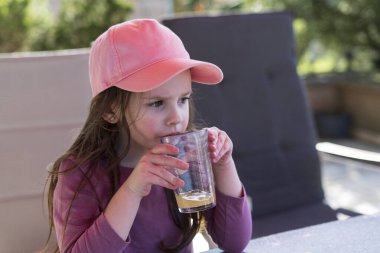 Beyzbol şapkalı küçük bir kız terasta bir masada oturuyor ve bardaktan meyve suyu içmekten hoşlanıyor. Mutlu çocukluk kavramı