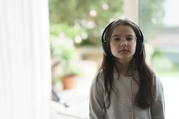 stock image A small long-haired girl with glasses is sad standing for the window wearing large headphones. The concept of musical education of children