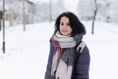 Genç, gülümseyen, güzel bir kadın kar yağarken açık havada fular takıyor.