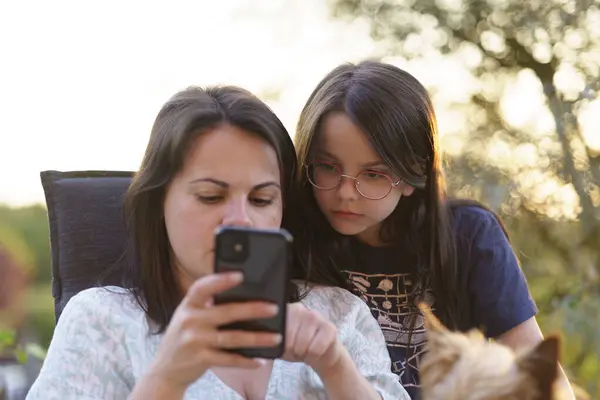 stock image A little girl in glasses with her mother on the terrace looking at a smartphone during sunset. The concept of a happy childhood and family