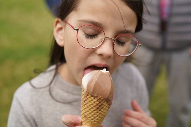 Gözlüklü küçük bir kız öğrenci yeşil çimlerin üzerinde dondurma külahı yiyor. Mutlu çocukluk kavramı