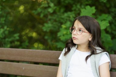 A little sad schoolgirl girl with glasses sits on a bench. Difficult childhood concept clipart