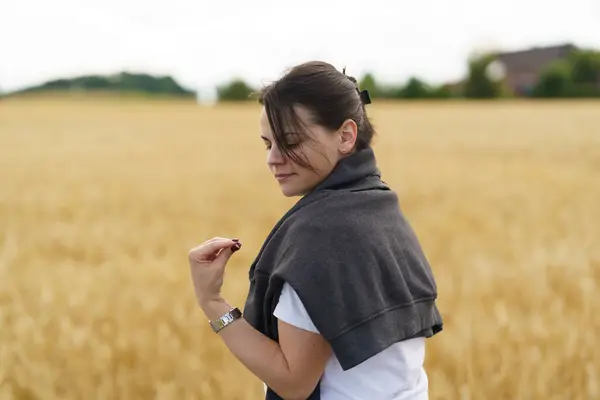 stock image A young smiling beautiful girl with collected hair and a sweater thrown over her shoulders against the backdrop of a field with ripe wheat. Rural travel or farming concept