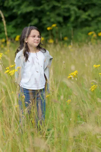 stock image A little teenage girl wearing glasses, jeans, a T-shirt and a sweater thrown over her shoulders stands in waist-deep natural grass. Little childish happiness in wildflowers.