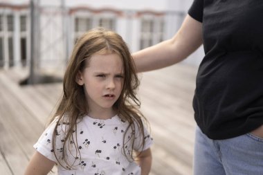 Ön dişi olmayan küçük, güzel bir kız öğrenci yazın, annesi tarafından kucaklanan bir tişörtle ahşap bir terasta düşünceli bir şekilde duruyor. Aile kavramı, çocukluk, bakım