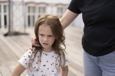 Ön dişi olmayan küçük, güzel bir kız öğrenci yazın, annesi tarafından kucaklanan bir tişörtle ahşap bir terasta düşünceli bir şekilde duruyor. Aile kavramı, çocukluk, bakım