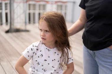 Ön dişi olmayan küçük, güzel bir kız öğrenci yazın, annesi tarafından kucaklanan bir tişörtle ahşap bir terasta düşünceli bir şekilde duruyor. Aile kavramı, çocukluk, bakım