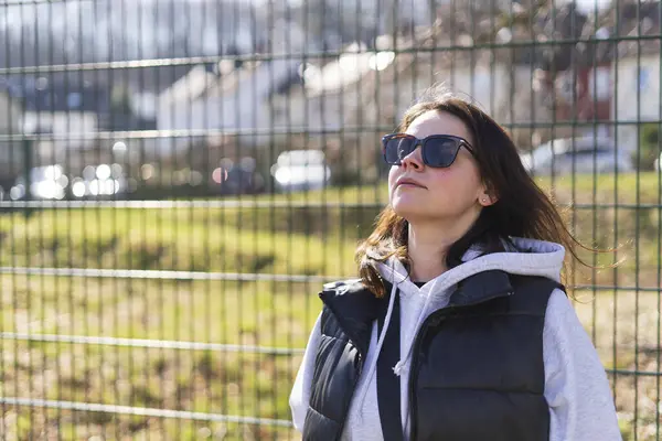 stock image A cute dark-haired girl in sunglasses, a vest and a hoodie stands smiling in a spring park in front of the fence