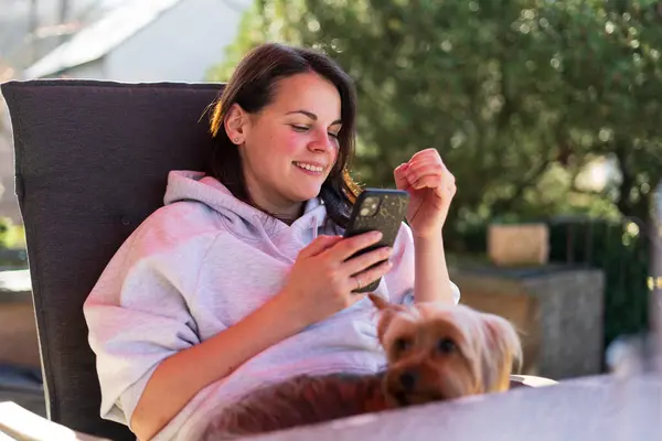 stock image A YOUNG beautiful girl in a light hoodie is reading on her mobile phone while sitting at a table on the terrace of her house. Cellular technology concept outdoors