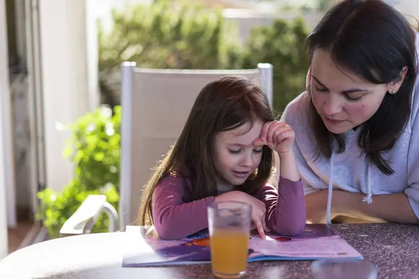 Stock image A cute little girl is reading a book with her beautiful mother while sitting at a table in the backyard on the terrace. Educational home family concept