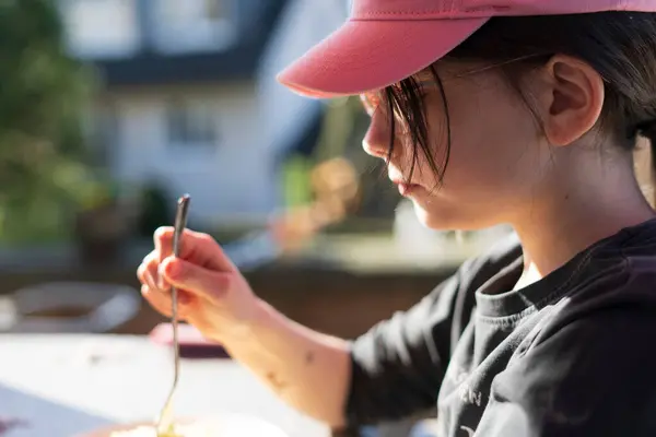 stock image A little cute girl in glasses and a baseball cap sits on the terrace of her house with a fork. Happy children's life concept